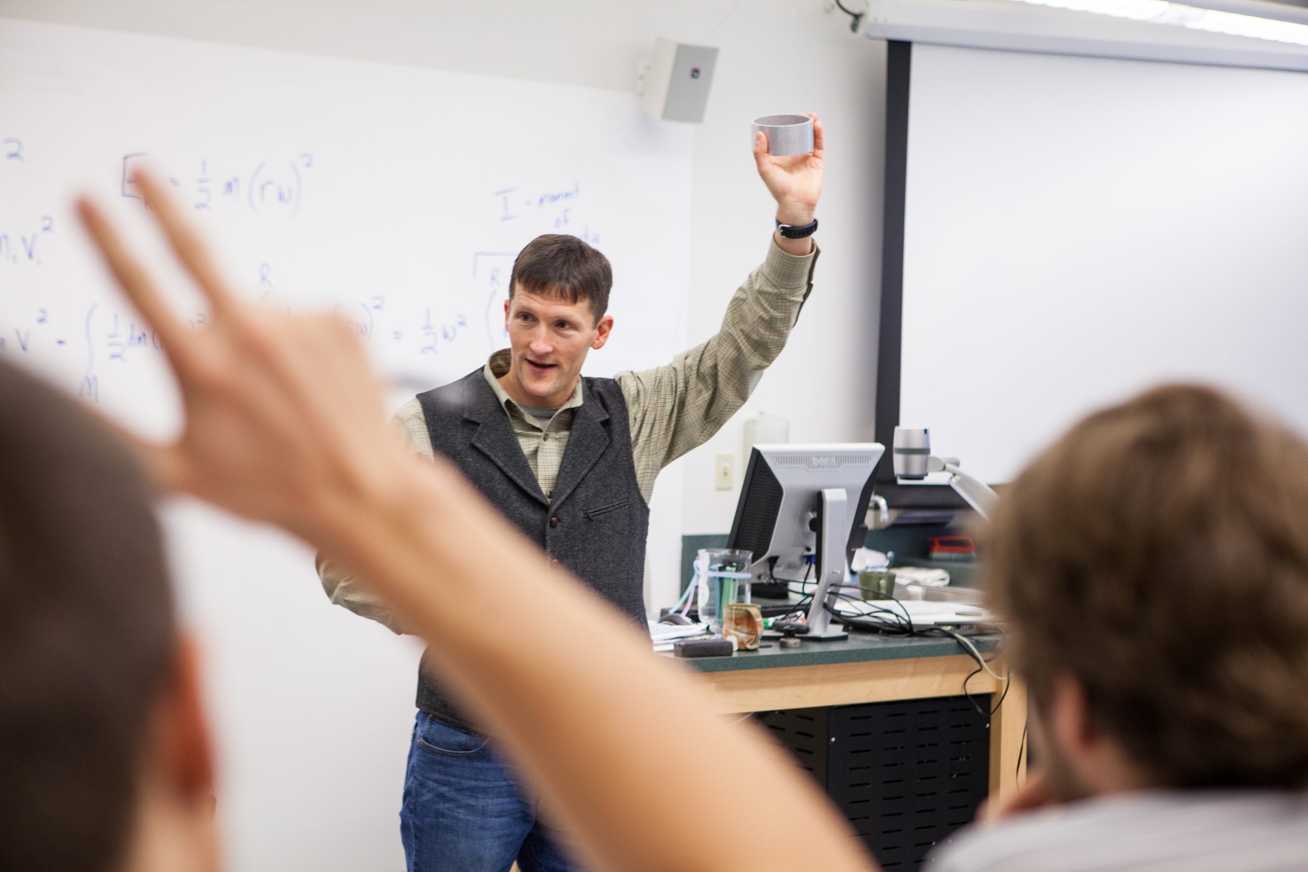 A professor stands with a ring raised above his head looking around the classroom. A student in the foreground raises their hand.