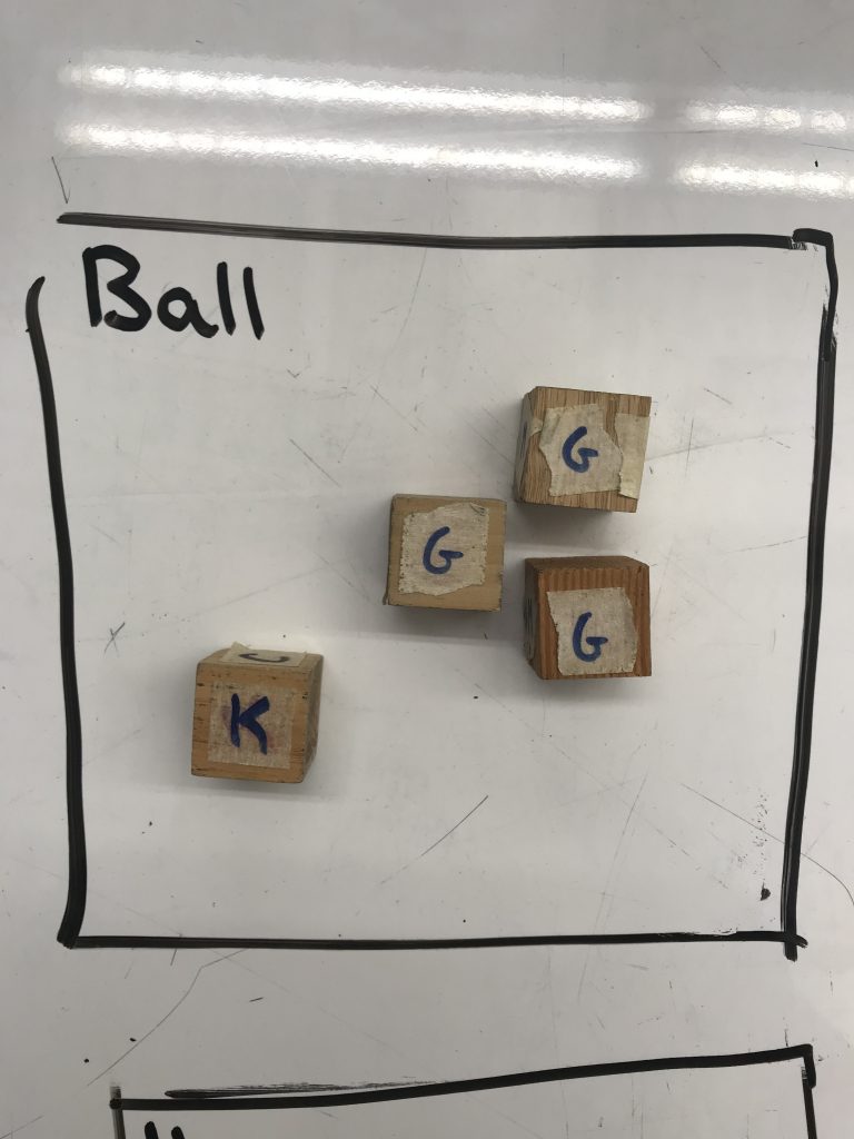 A black box labeled Ball is written on a whiteboard. Four wooden cubes sit inside. One has a K on it, the other three have G on them.